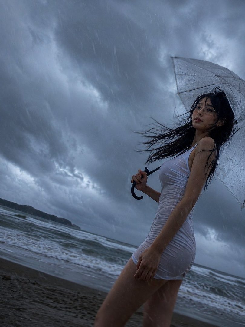A young woman with long black hair and glasses in a wet white dress holding a transparent umbrella on a stormy rainy beach.