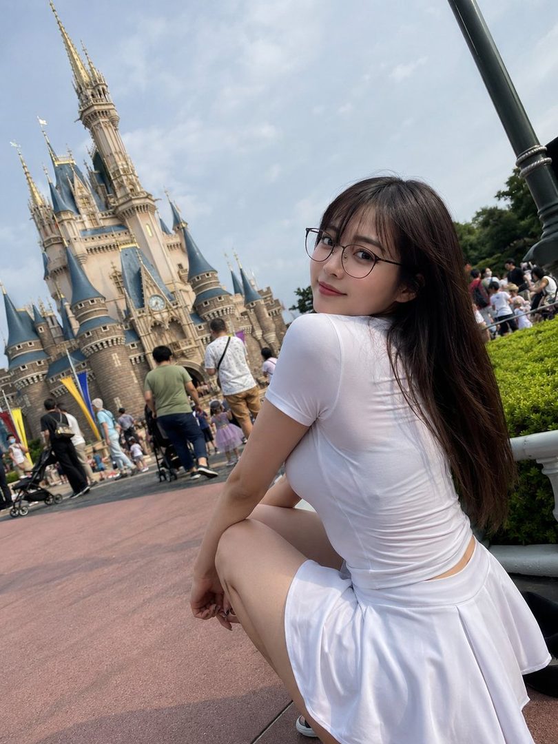 Young woman with long dark hair and glasses in a white outfit posing outdoors in front of a castle at a busy theme park.
