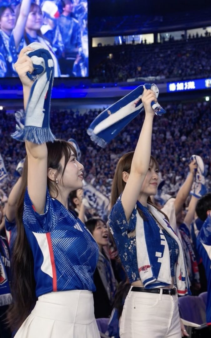 Two young women in blue and white sports jerseys cheer enthusiastically at a night soccer game, holding up scarves.