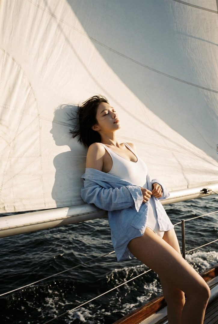 A woman in a white swimsuit and blue shirt leans against a sailboat sail, with her eyes closed and face towards the sun.