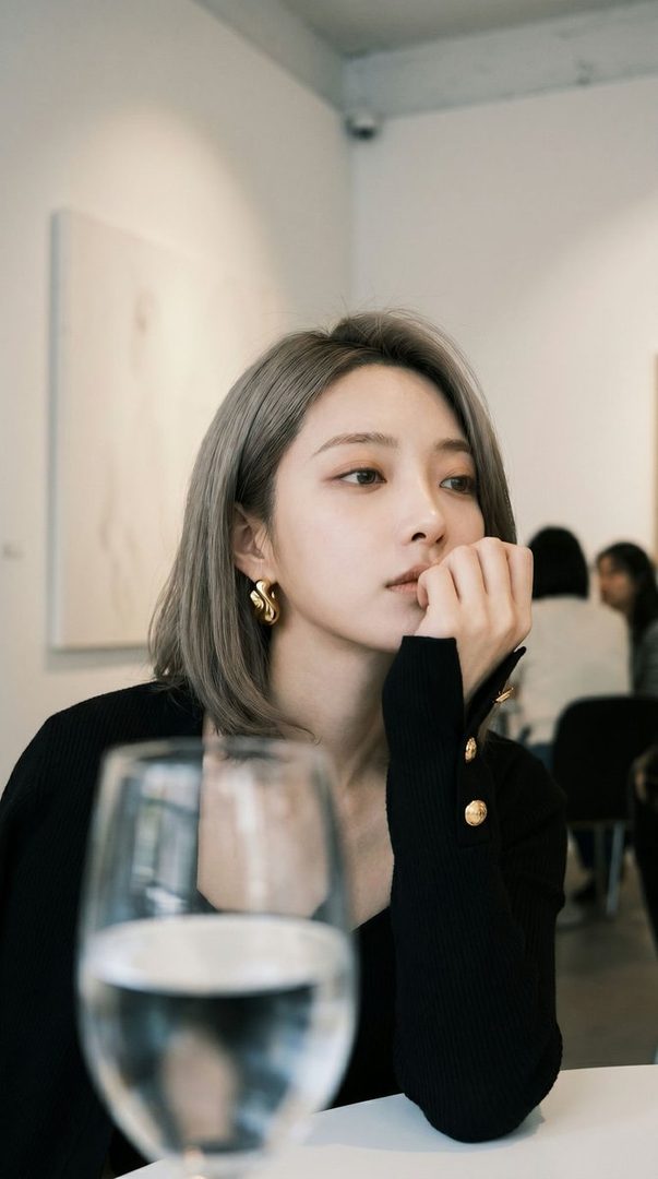 A woman with short silver hair and a black top sits at a table with a glass of water in the foreground, looking thoughtfully to the side.