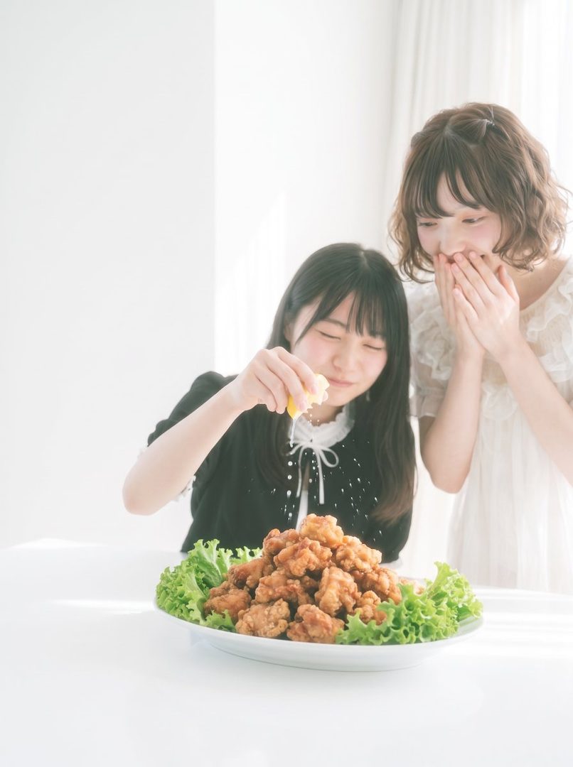Two young women are sitting at a table with a large plate of fried chicken, one is squeezing a lemon over it while the other watches with a smile.