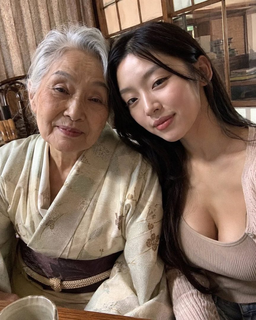 A young woman with long black hair takes a selfie with an elderly woman wearing a kimono, both smiling gently.