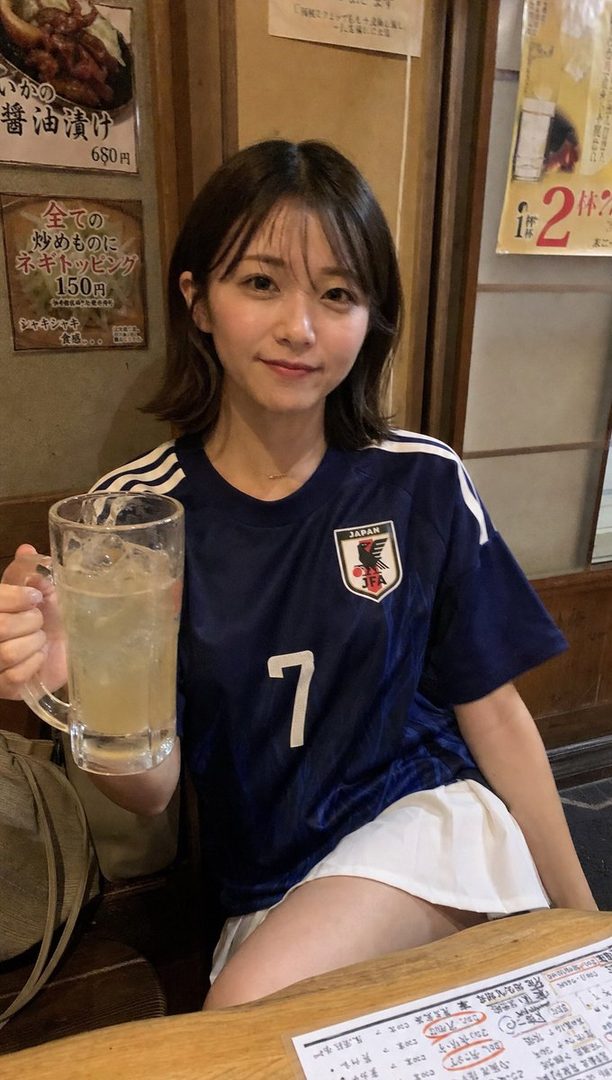 A young woman with short brown hair, wearing a blue Japan soccer jersey and a white skirt, holds a glass of iced drink while sitting at a table indoors.
