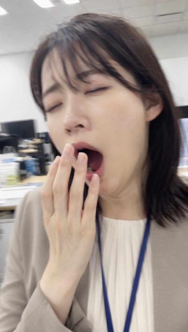 A woman with brown hair and bangs yawns widely, covering her mouth with her hand, in an office setting.