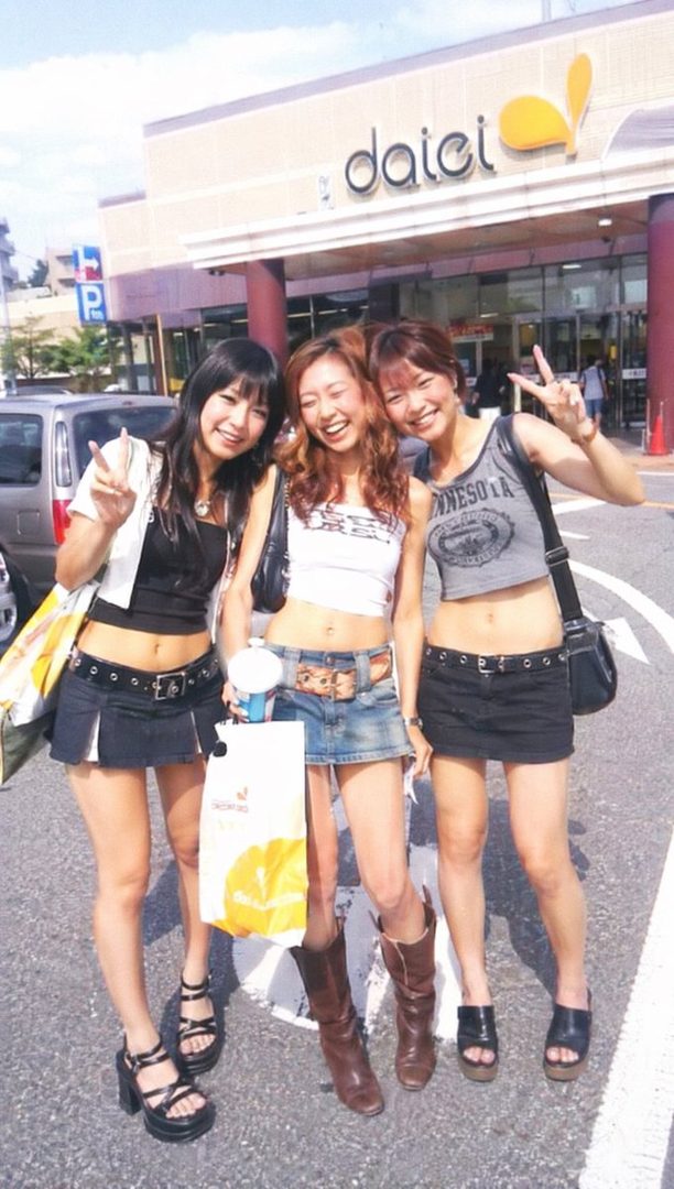 Three young women pose happily in front of a Daiei store on a sunny day.