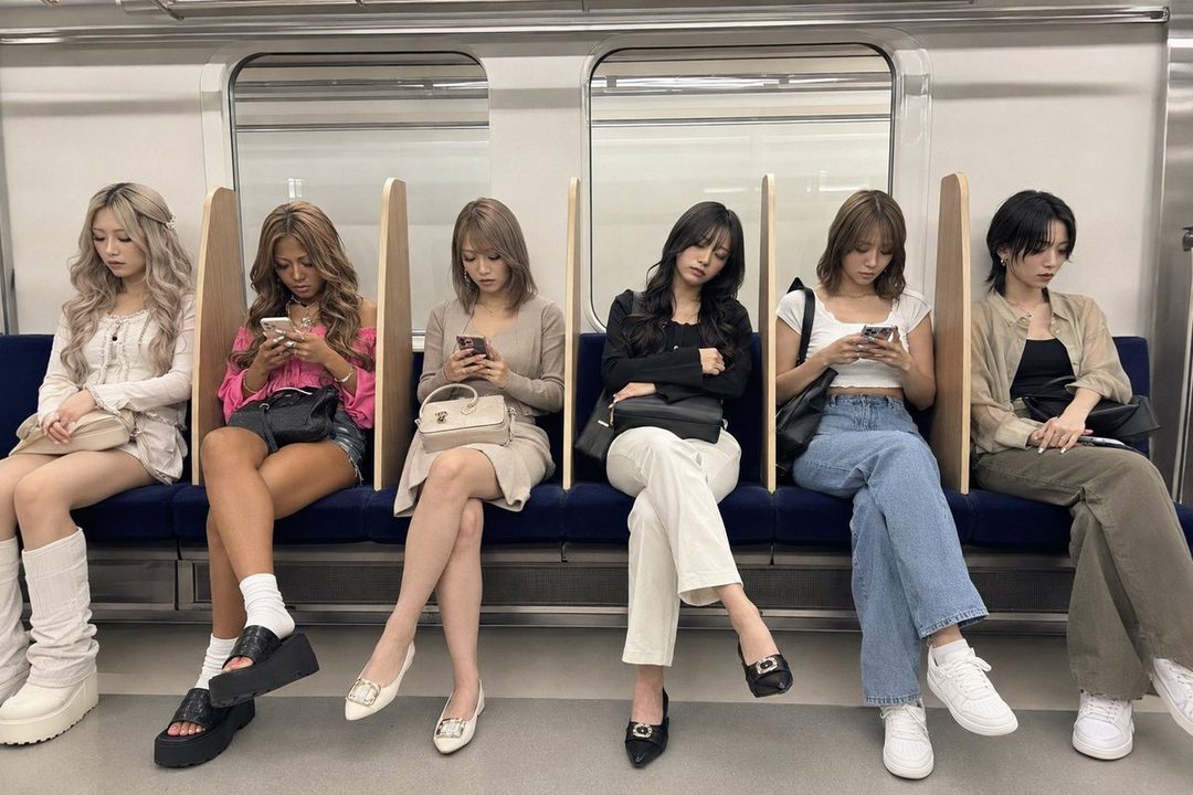 Six young women with various hair colors and casual outfits are sitting on a train, looking at their phones.