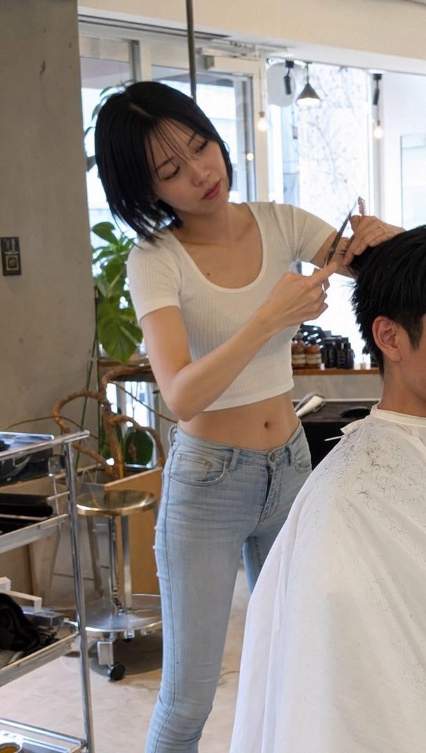 A woman with short black hair, wearing a white crop top and jeans, is cutting a man's hair in a bright salon.