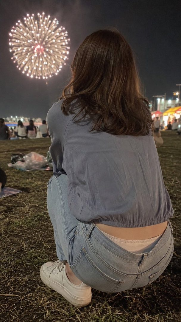 A person with long brown hair, wearing a blue top and jeans, squats on the grass watching fireworks at night.