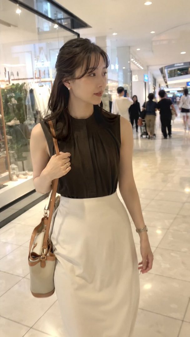 A young woman with long brown hair, wearing a dark sleeveless top and a white skirt, walks through a brightly lit indoor shopping mall.