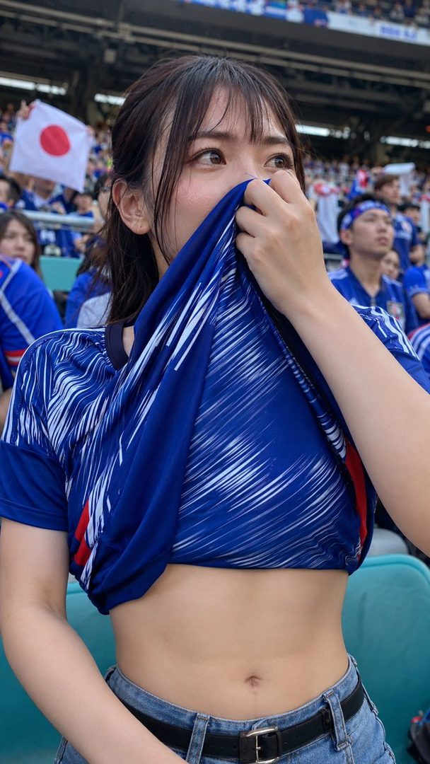 A young woman in a blue and white sports jersey covers her mouth with her shirt while watching a game in a stadium.