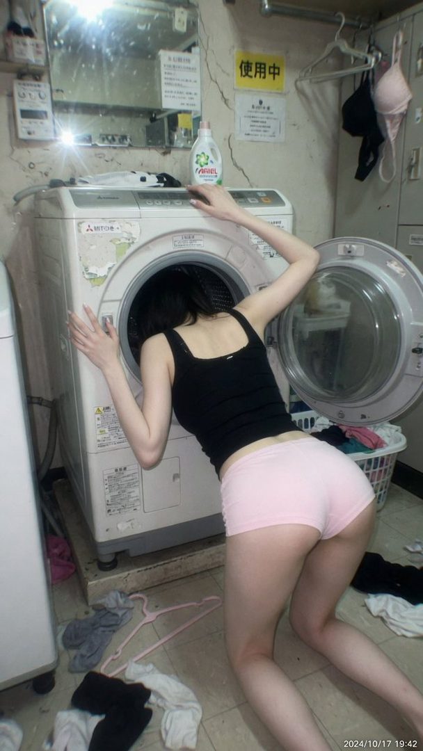 A woman in a black tank top and pink shorts leans into a washing machine in a messy laundry room.