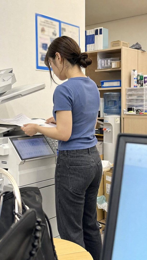 A woman with brown hair tied in a bun, wearing a blue top and jeans, is operating a copier in an office setting.