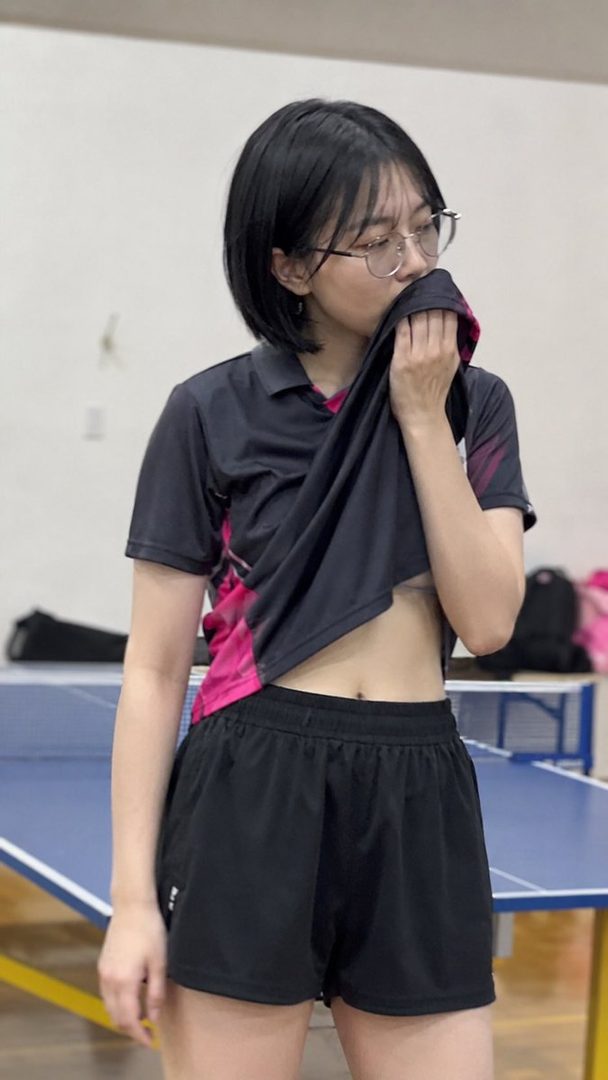 A young woman with short black hair and glasses wipes her face with a shirt, standing next to a ping pong table.