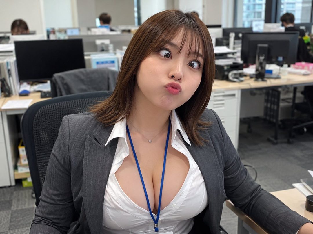 A playful woman in an office setting makes a funny face at the camera, wearing a white shirt and a gray suit jacket.