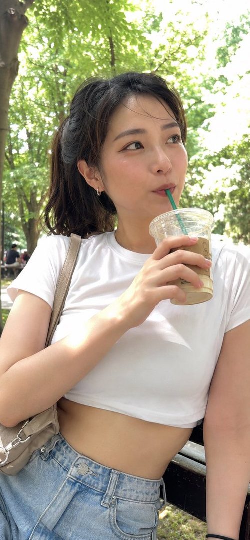 A young woman with brown hair in a ponytail sips a drink outdoors on a sunny day.