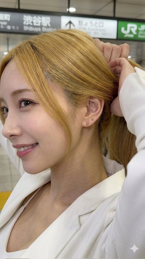 A woman with blonde hair smiles while tying her hair back, wearing a white top in what appears to be a train station.