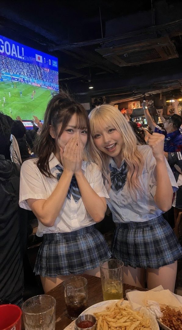 Two young women in school-style uniforms are smiling and posing for a photo in a bar, with a soccer game on a large screen in the background.