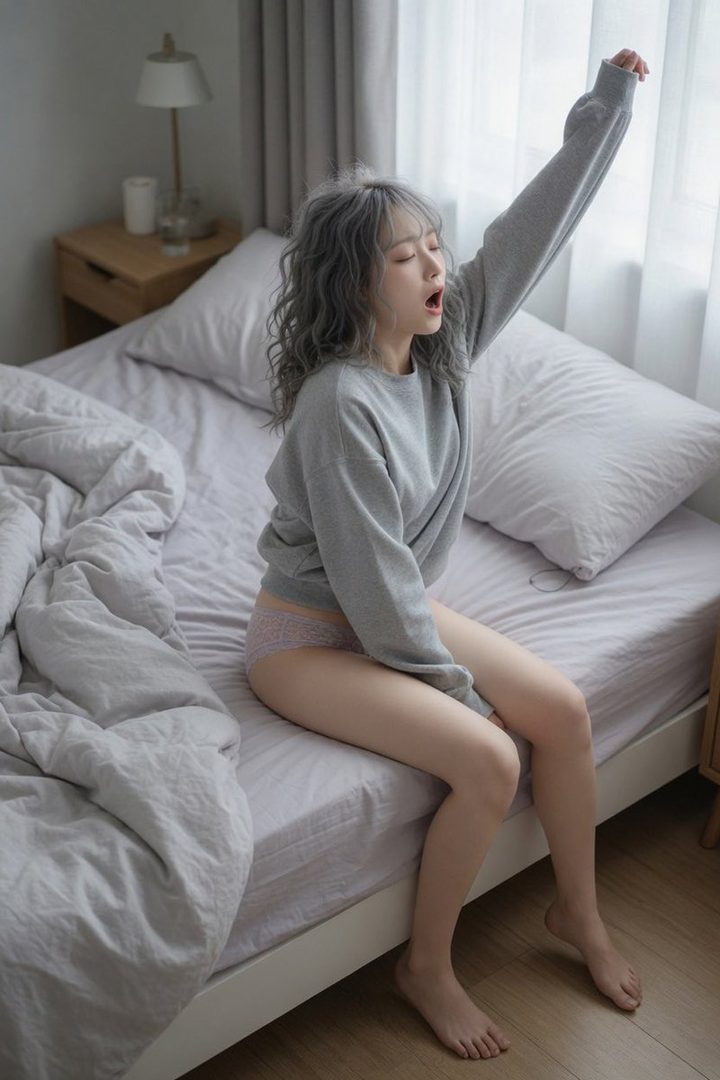 A young woman with grey curly hair, wearing a grey sweatshirt and light pink panties, yawns and stretches while sitting on a bed in a brightly lit bedroom.