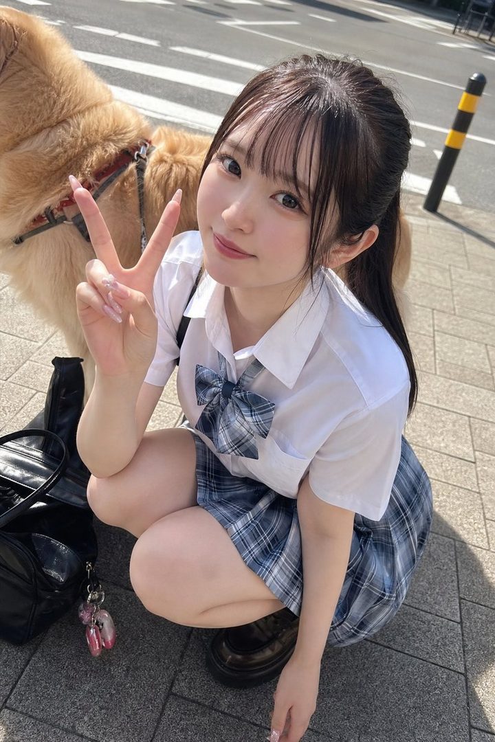 A cute girl in a school uniform squats on a sunny street, making a peace sign with her hand.