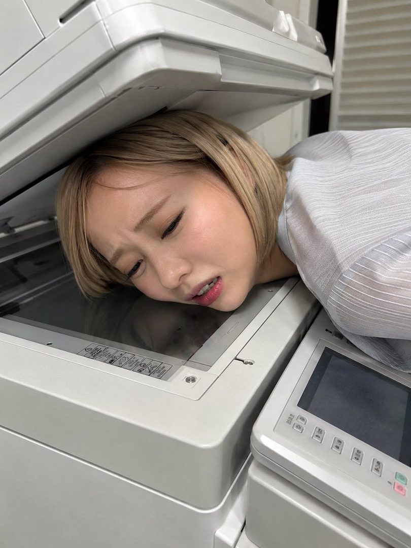 A young woman with blonde hair is playfully squashed between two office copiers, making a funny face.