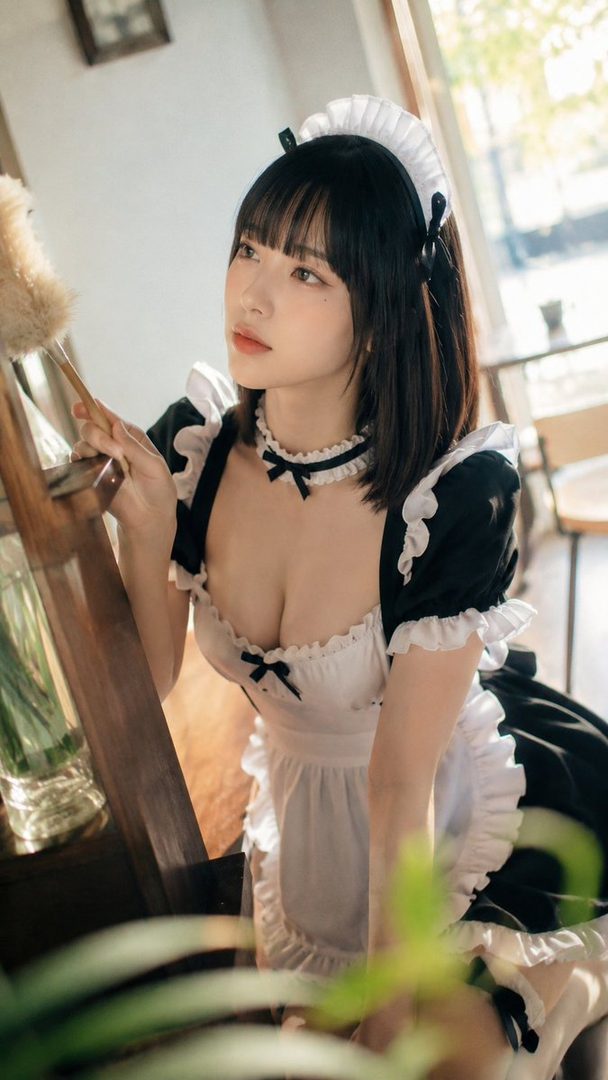 A young woman with black hair and bangs, dressed in a maid outfit, holds a duster indoors.