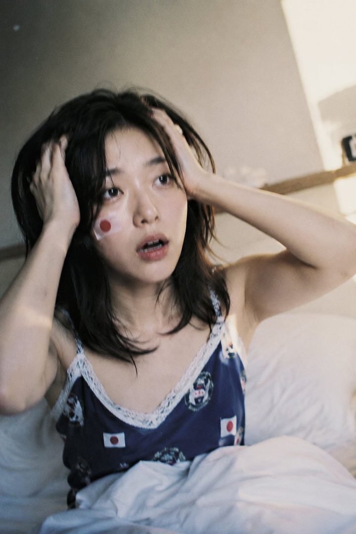 A young woman with dark hair and a Japanese flag sticker on her cheek, wearing a camisole, looks surprised while sitting in bed.