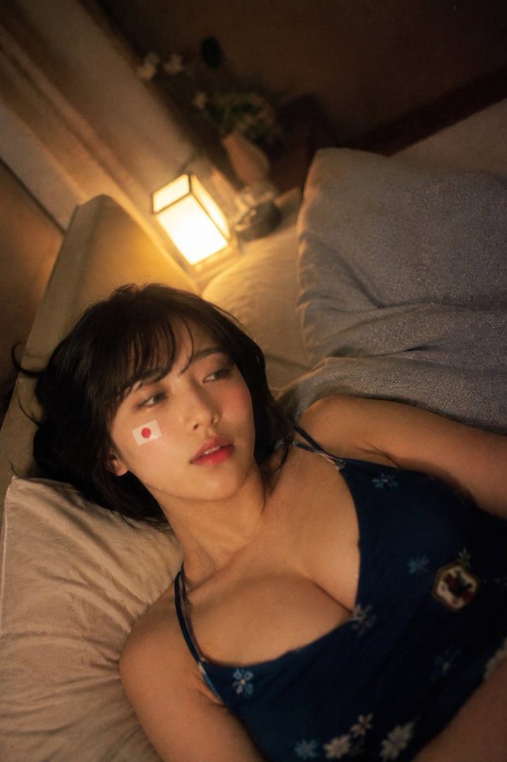 A woman with a Japanese flag sticker on her cheek lies on a bed in a dimly lit room.