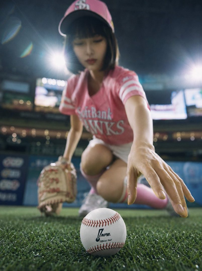 A woman in a pink baseball uniform and cap crouches on a baseball field at night, reaching for a baseball in the foreground.