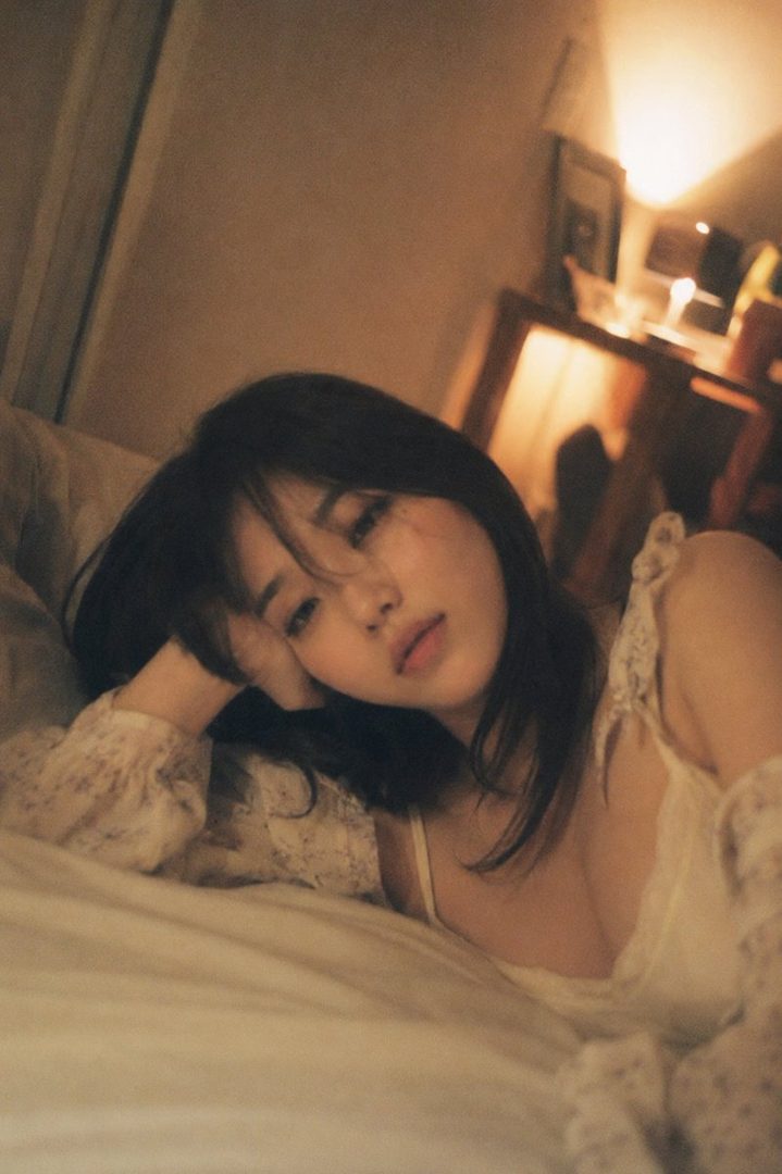 A dreamy portrait of a young woman with dark hair lying on a bed in a dimly lit room, wearing a light-colored top.