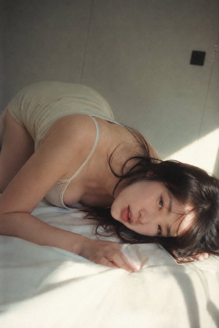 A woman with long dark hair is lying on a white bed in a suggestive pose, looking towards the viewer.