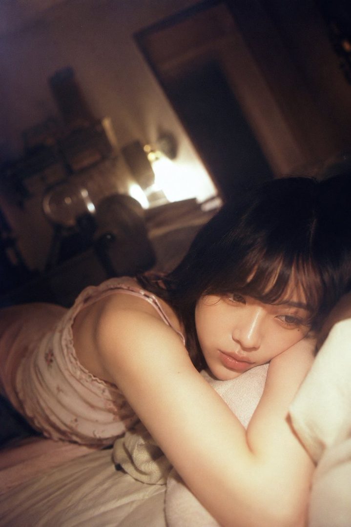 A young woman with dark hair and bangs lies on a bed in a dimly lit room, looking pensive.
