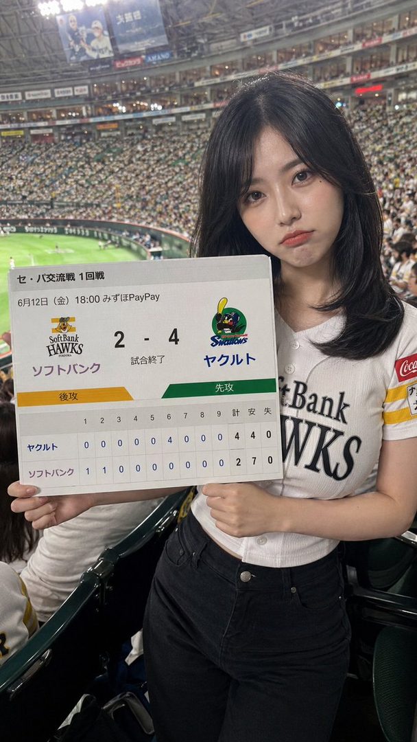 A young woman with black hair in a baseball jersey holds up a scoreboard at an indoor stadium.