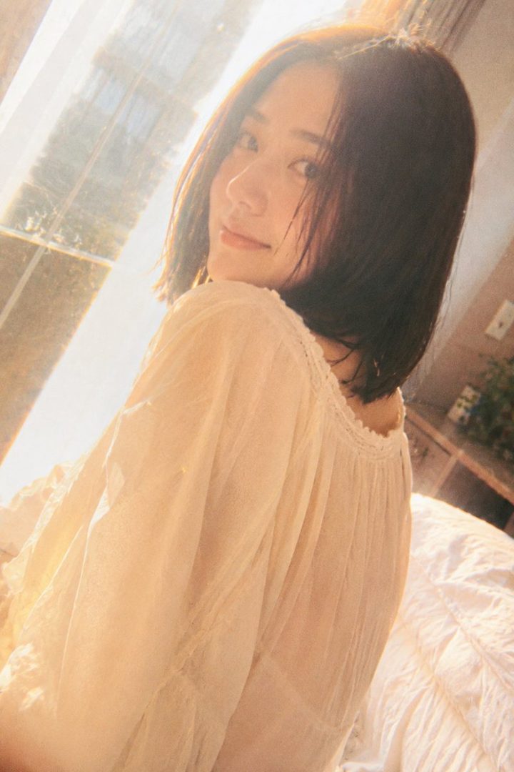 A photorealistic portrait of a woman with short black hair, wearing a light-colored casual top, looking over her shoulder with soft sunlight in an indoor setting.