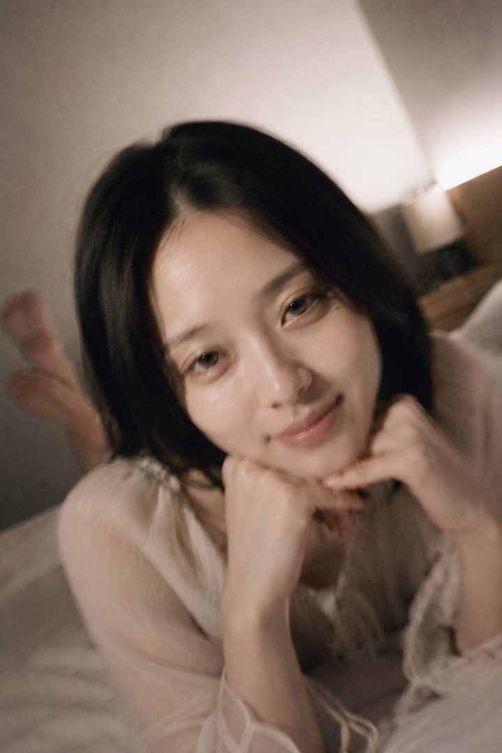 A photorealistic portrait of a young woman with black hair, lying on a bed and resting her chin on her hands, looking serenely at the camera.
