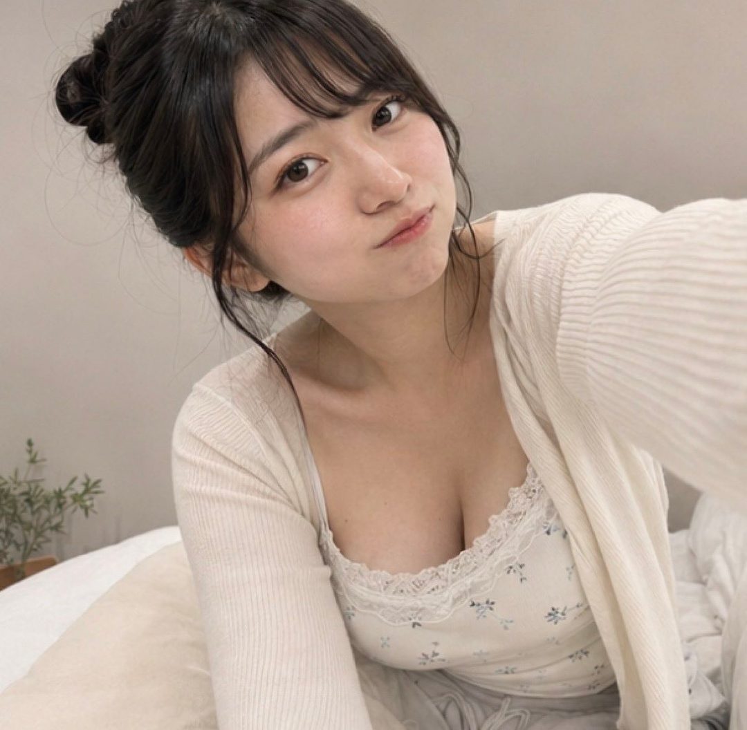 A young woman with black hair in a bun and bangs takes a selfie indoors, wearing a white lace-trimmed top and cardigan.