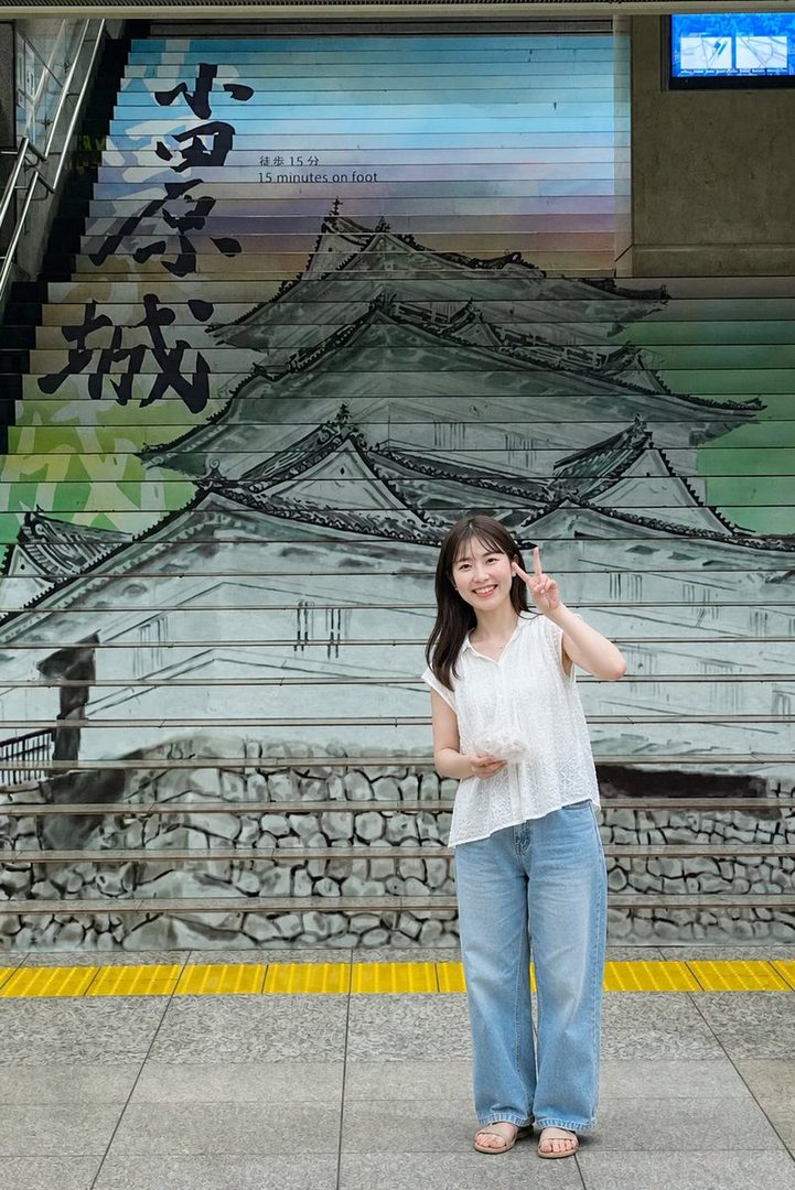 A young woman stands in front of a staircase painted with a Japanese castle, making a peace sign.