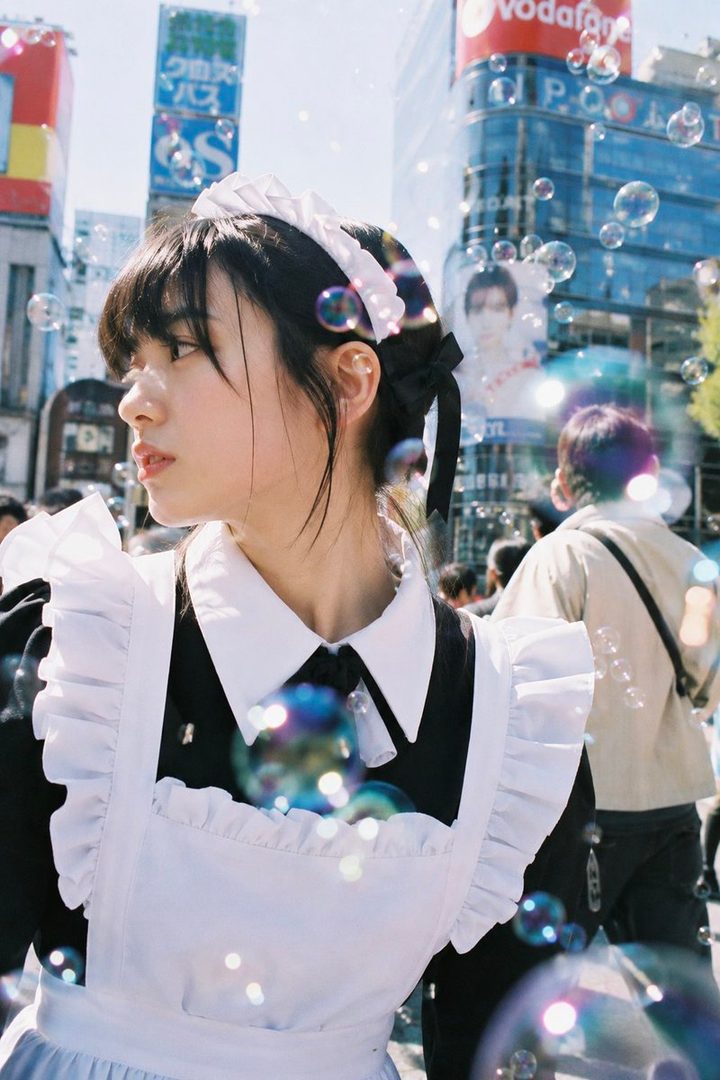 A young woman in a maid outfit stands on a city street with bubbles floating around her.
