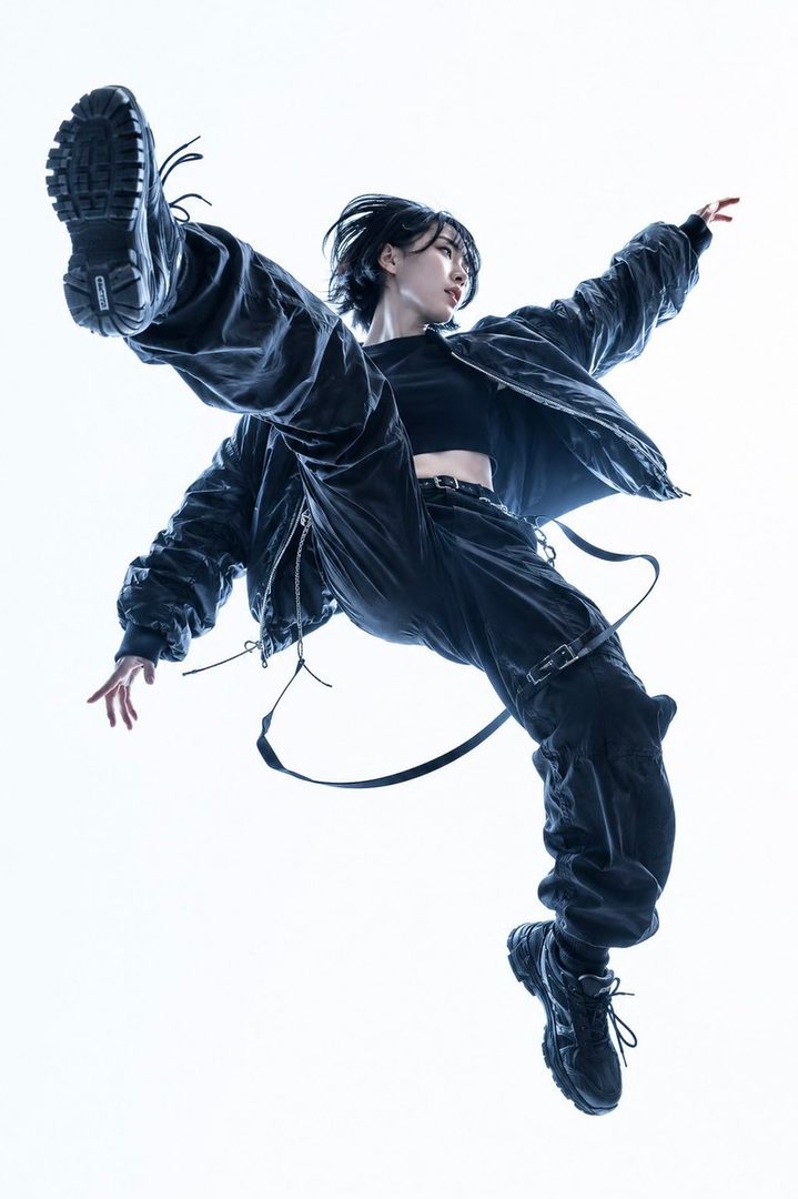 A dynamic studio shot of a woman with short black hair wearing a black crop top, jacket, and cargo pants, captured mid-jump against a white background.