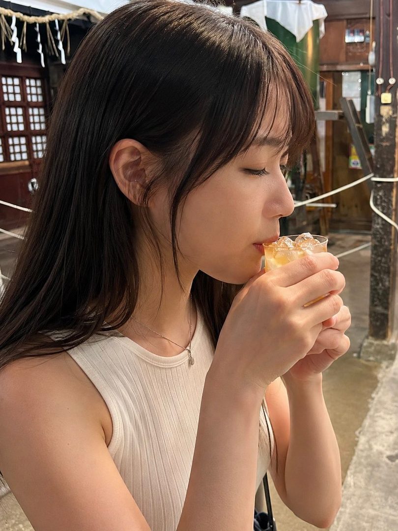A young woman with long brown hair and bangs sips a drink outdoors on a sunny day.