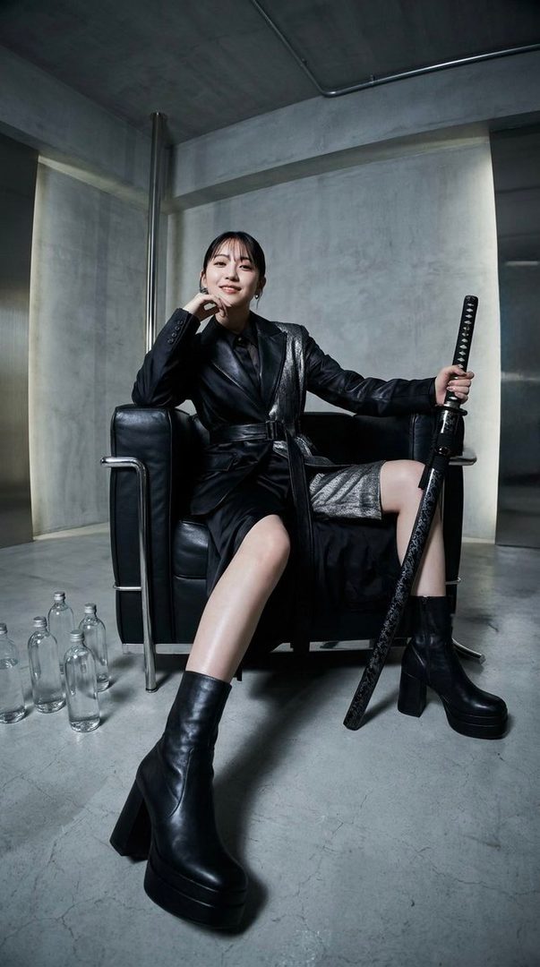 A woman with black hair and bangs, wearing a black suit and boots, sits in a chair holding a katana in an indoor setting.