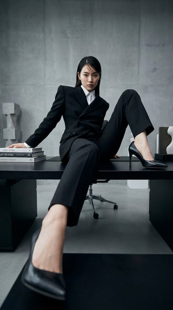 A woman in a black suit and white shirt sits on a desk in an office setting, looking directly at the camera.