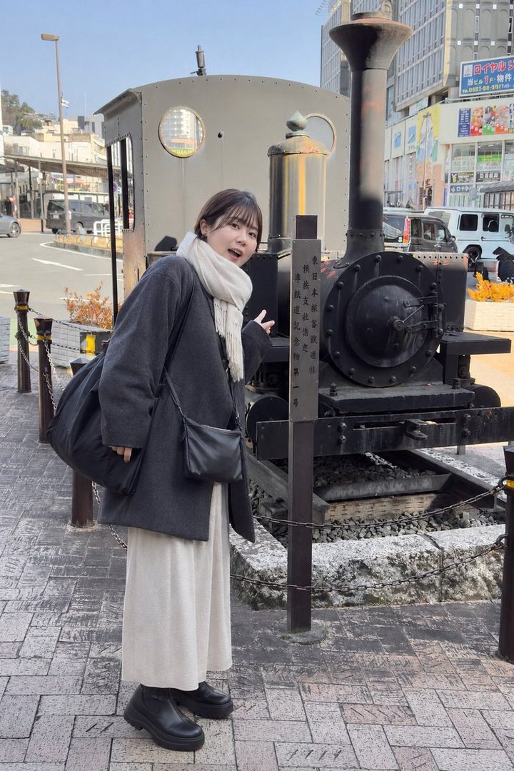 A young woman in a grey coat and white scarf stands next to an old train engine on a city street.