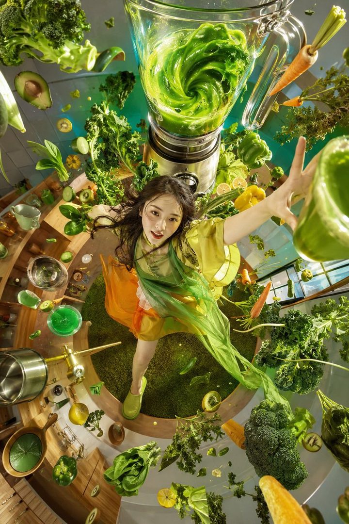 A young woman in a green and orange outfit is surrounded by floating green vegetables and a blender with green liquid, viewed from above.