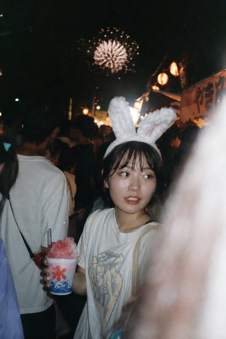 A young woman with bunny ears and bangs holds a shaved ice drink at a night festival with fireworks in the background.
