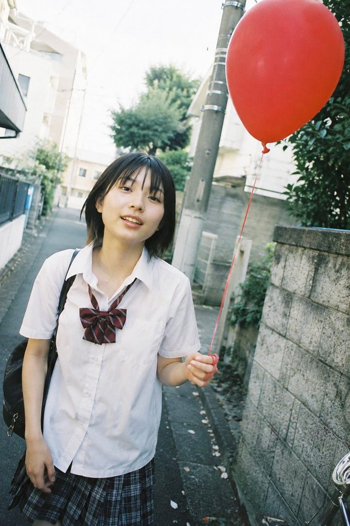 A young girl with short black hair and bangs, wearing a school uniform, smiles while holding a red balloon on a narrow street.