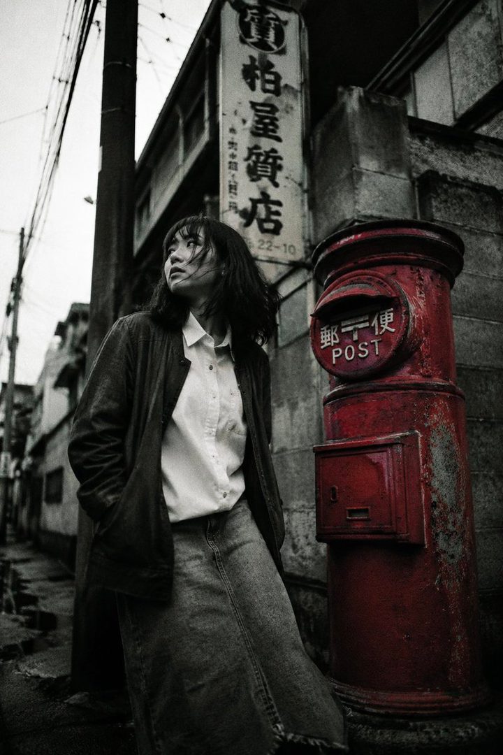 A young woman with black hair and a casual outfit stands next to a red post box on a narrow street, looking up and to the side.