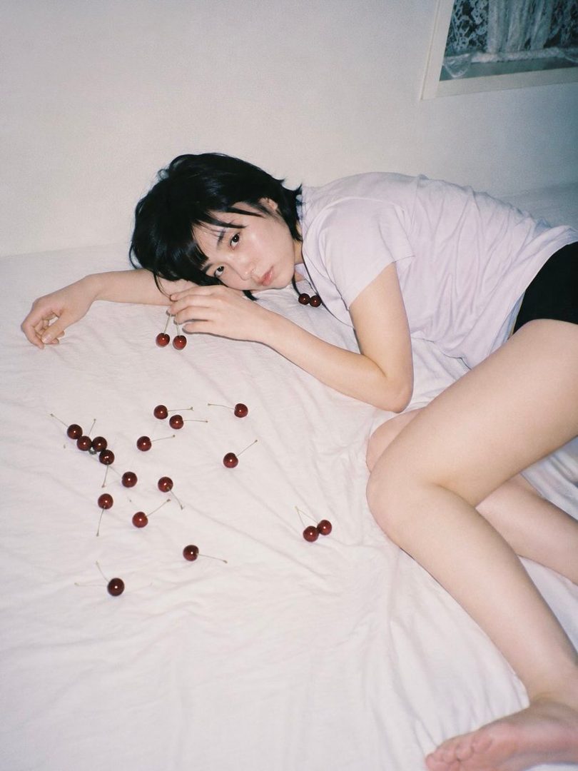 A young woman with short black hair lies on a white bed with cherries scattered around her.