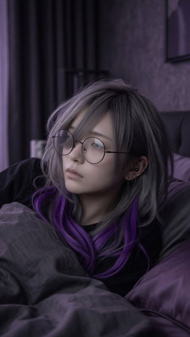 A person with silver and purple hair and round glasses looks thoughtfully to the side while lying in bed.