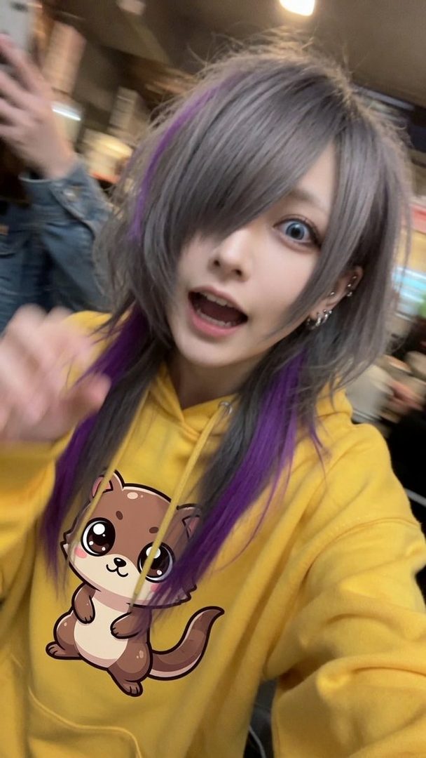 A playful person with silver and purple hair wearing a yellow hoodie with a cute otter graphic.